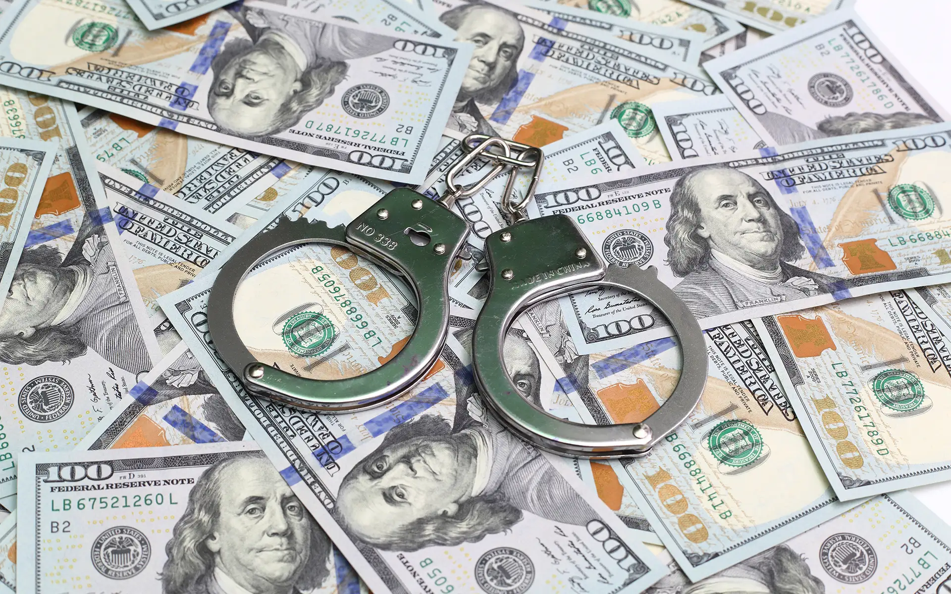 One hundred-dollar bills are scattered across a table with a pair of handcuffs resting on top
