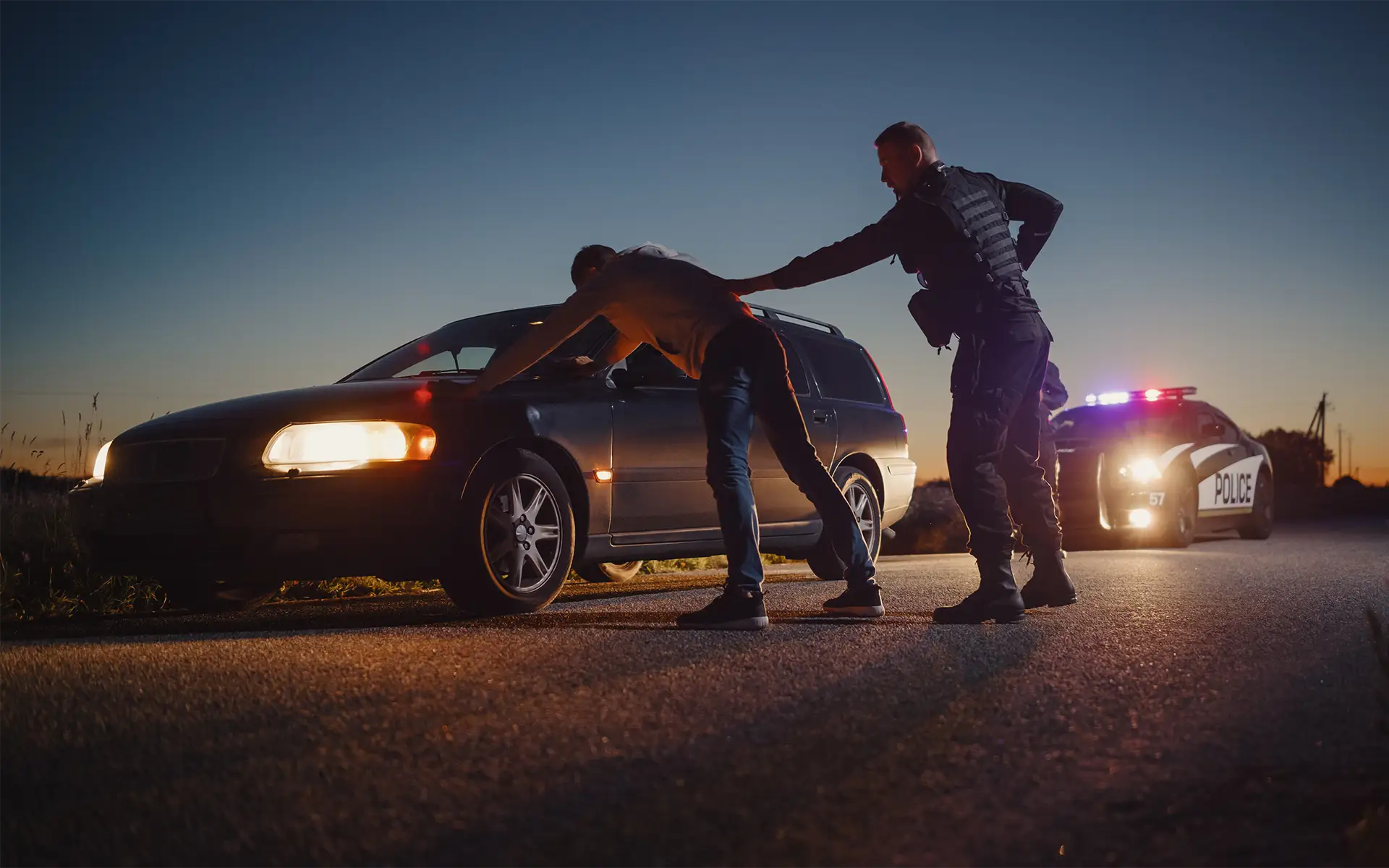 A police officer patting down a man with his hands on a stopped car on the side of the road