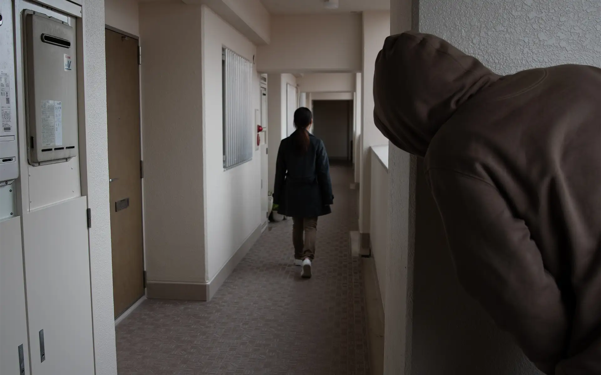 The back view of a woman walking down a hallway with a stalker in a hoodie peering around the corner at her